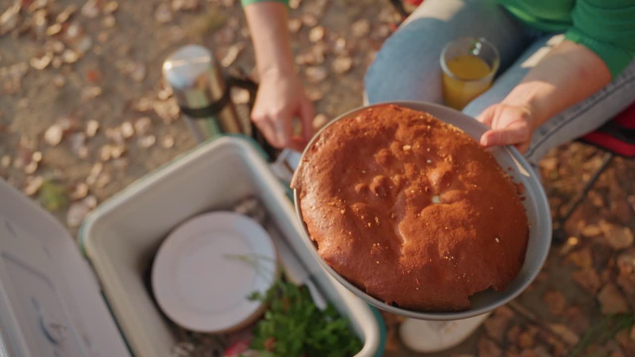 Campingkake på kjølebokslokket, campist i grønn genser som tilbyr gyllen kake fra form over åpen kjøleboks, tallerkener og salat synlig inni, spredte høstblader under føttene, sollys som fremhever skorpen.