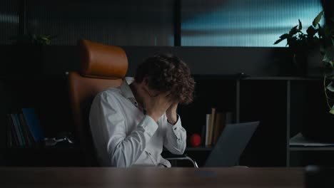 Side-view-of-a-tired-guy-with-curly-hair-in-a-white-shirt-covers-his-face-with-his-hands-and-shakes-his-head-during-a-hard-day-at-work-in-the-office