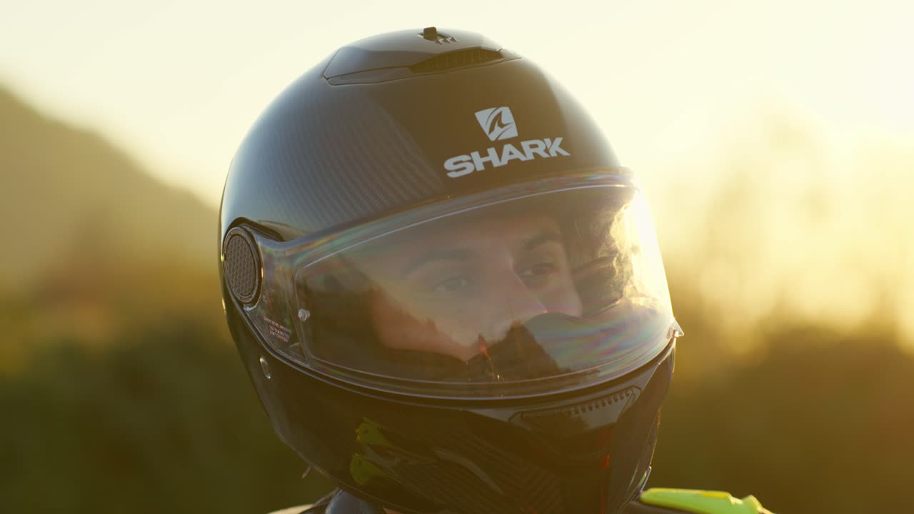 Motorcyclist wearing a helmet stands outside at sunset, with cars visible in the background, close up shot in slowmotion