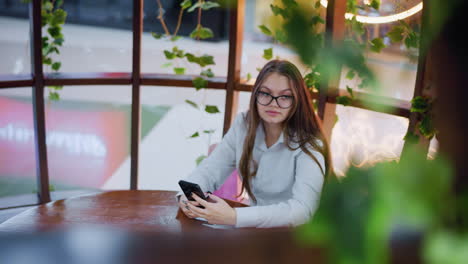 young lady seated at cafe table holding phone while looking thoughtfully at something, surrounded by greenery and vibrant atmosphere, creating a relaxed and focused vibe