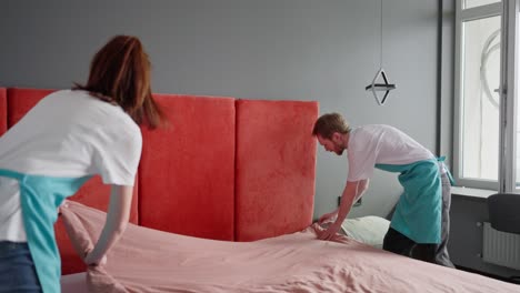 Confident-group-of-two-cleaners-a-blond-guy-and-a-brunette-girl-in-a-white-T-shirt-and-blue-aprons-make-the-bed-with-a-pink-blanket-during-cleaning-in-the-bedroom