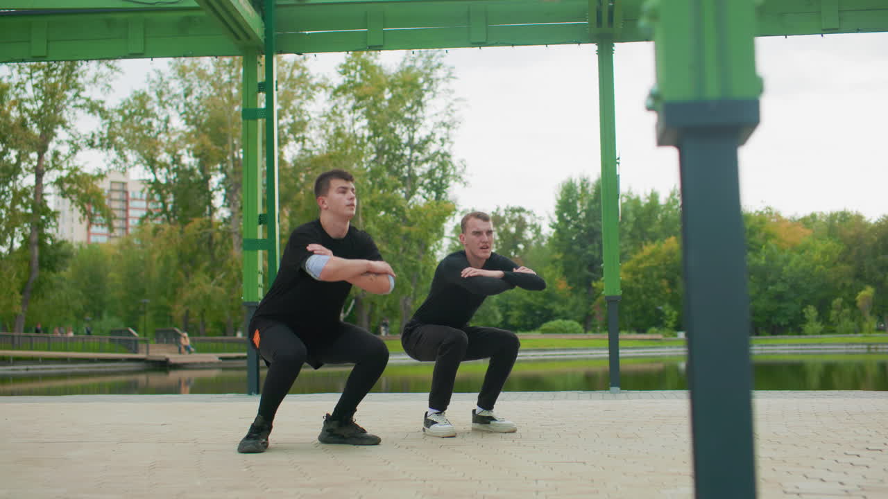 Due uomini bianchi si allenano sotto un gazebo verde, ripetizioni di squat sincronizzate ed energiche, lago del parco sullo sfondo, espressioni concentrate, abbigliamento sportivo nero, sneakers, luce del sole mattutina, cameratismo determinato