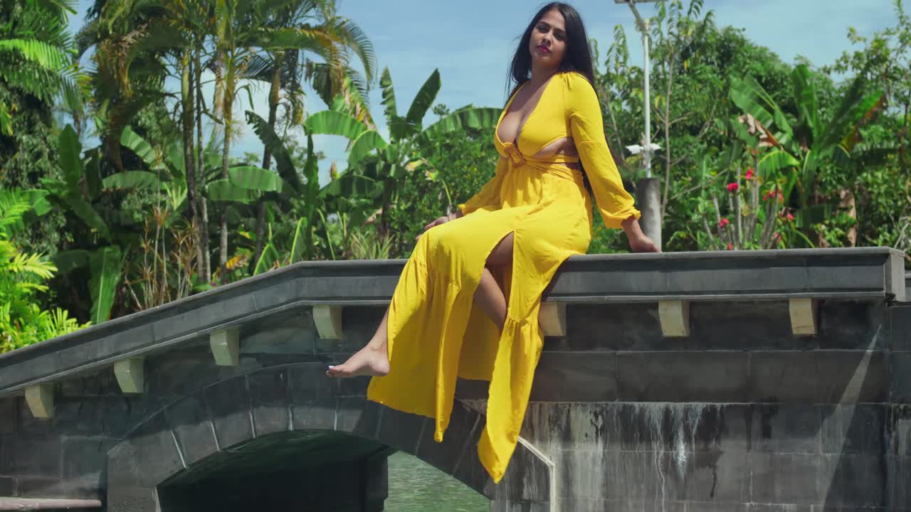 молодая женщина в ярко-желтом платье сидит на мосту роскошного замка с бассейном на заднем плане