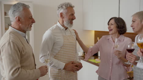 group of cheerful senior friends talking and laughing while drinking wine in the kitchen