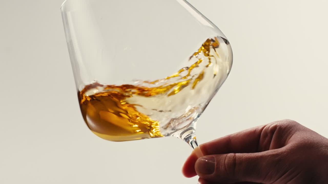 Close-up shot of hand holding a wine glass with amber liquid swirling inside against light background.