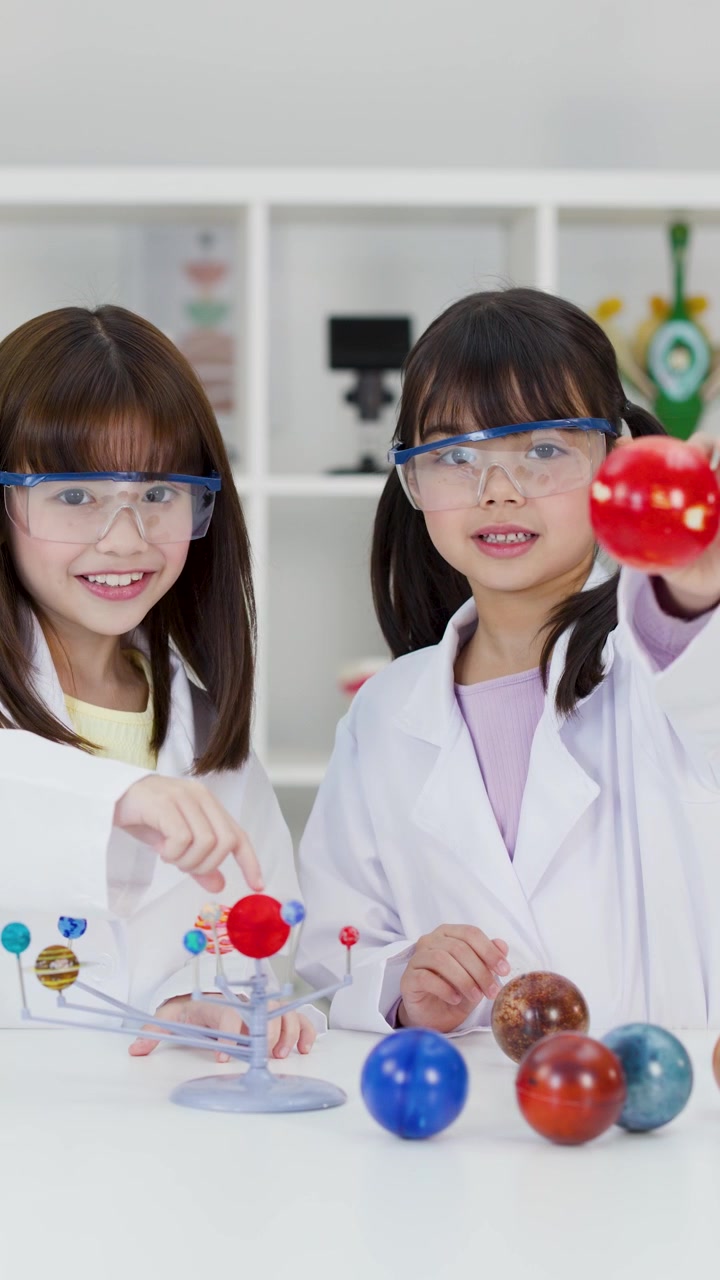 Dua gadis muda mengenakan kacamata pengaman dan jas lab memeriksa model planet di ruang kelas