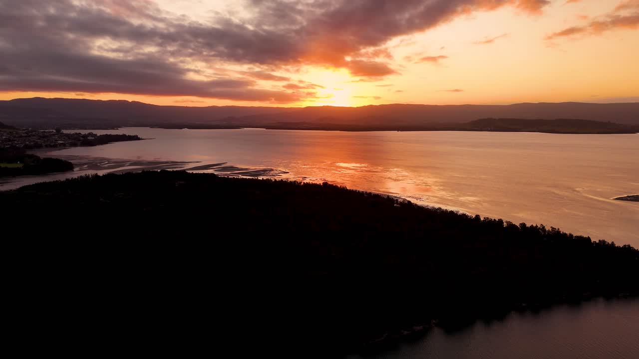 Sunset at Lake Illawarra casting a warm glow over the water, framed by tranquil coastal scenery, aerial ascend over silhouette land