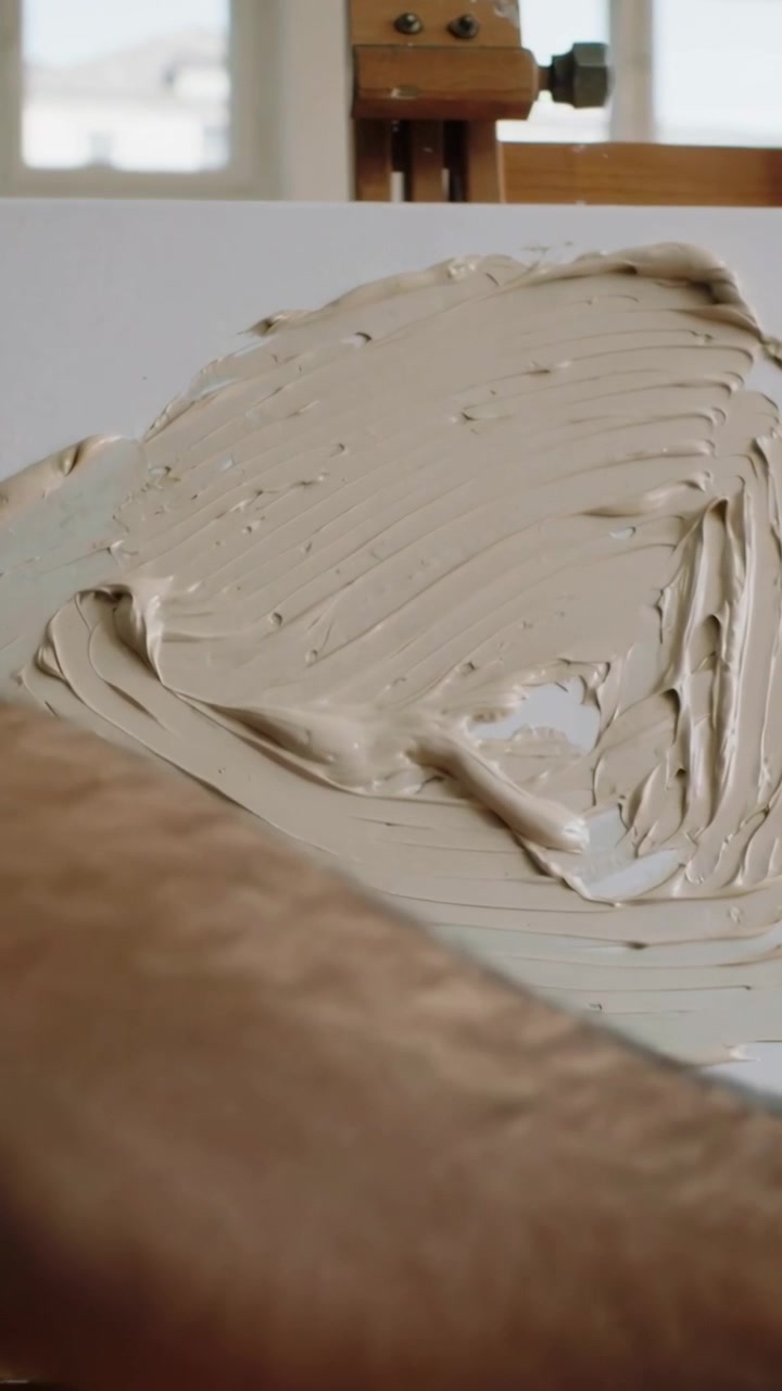 Vertical video: Bare arm entering left, studio artist smoothing beige with knife, priming canvas