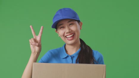 푸른 유니폼을 입은 아시아 여성 택배사의 클로즈업은 스튜디오에서 녹색 스크린 배경에 상자를 배달하는 동안 몸짓 평화와 미소를 짓고 있습니다.