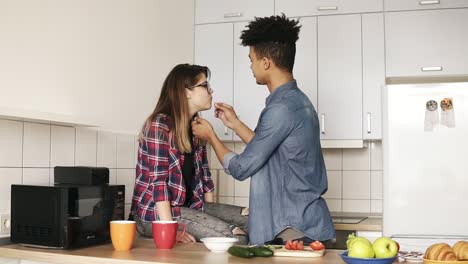 Zwei-Junge-Leute-Genießen-Ihre-Gemeinsame-Zeit-Und-Bereiten-In-Der-Gemütlichen-Küche-Das-Mittagessen-Zu.-Zusammen-Leben.-Mulatte-Füttert-Seine-Attraktive-Kaukasische-Freundin-Mit-Einer-Tomatenscheibe.