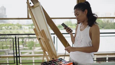 a woman holding a smartphone and paintbrush while painting in an art studio