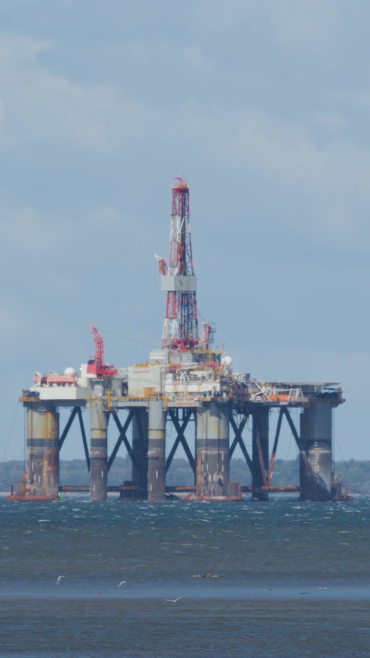 Stor offshore olieplatform står i oprørt hav under overskyet dagslys, set fra kysten med minimal kamerabevægelse og fjern horisont