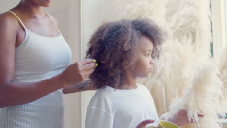 cute black girl standing by the window and drinking tea while mom making her hair