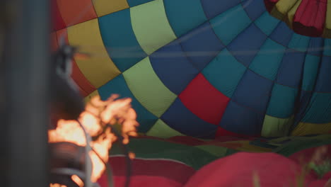 woman grips hot air balloon fabric panel beside roaring burner flame during sunset preflight setup in open grassy field with vibrant checkered envelope inflating under gentle breeze under soft sky