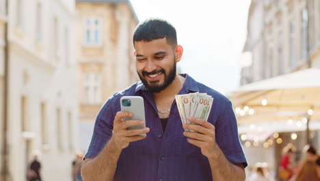 happy young man counting money dollar cash, use smartphone calculator app in urban city street