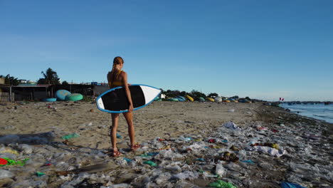 athletic surfer girl with surfboard strolls on beach polluted with plastics