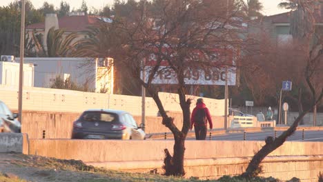 Rear-view-of-a-young-man-with-a-bag-on-his-back-walking-along-the-road-in-an-urban-area-at-sunrise