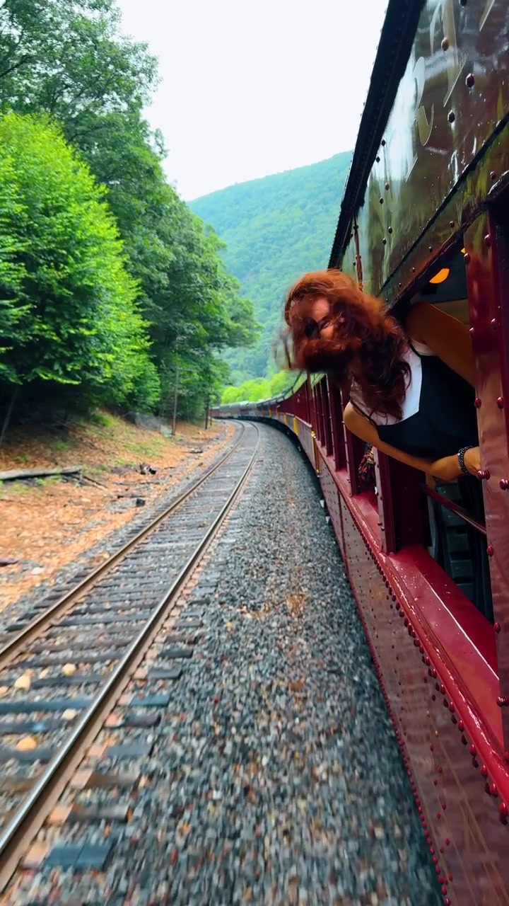 Donna con i capelli al vento che si sporge dal finestrino di un treno in movimento. Donna dai capelli rossi che si affaccia dal finestrino di un vagone rosso in movimento, con i capelli scompigliati dal vento lungo una panoramica ferrovia di montagna.