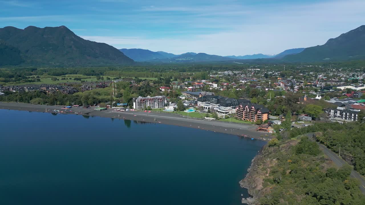 Orbiting drone view of Pucón’s beach and town, with calm lake waters, hotels, and scenic mountains surrounding the area