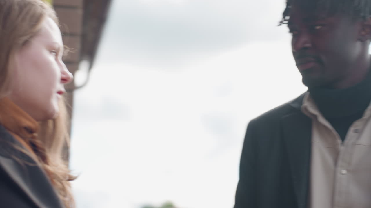 American lady in black coat checks phone, shows screen to friend, he nods, both start running along covered walkway with metal rails and overcast daylight, urban entrance mood, calm motion captured