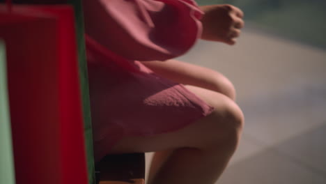 una bolsa de compras es dejada caer en un banco de madera marrón en un centro comercial por una dama con un vestido rosado sentada, cruzando la pierna y colocando la mano en su pierna, la pierна y la bolsa son parcialmente visibles