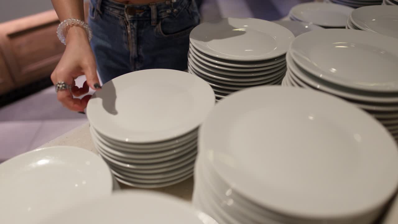 Close-up of woman picking clean white plates from stacked buffet table in warm lighting