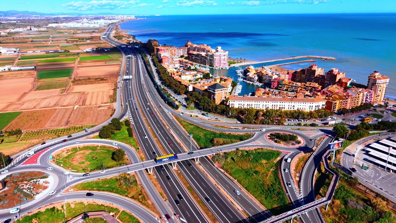 Vista aerea di una città costiera mediterranea e di un porto turistico, Spagna. Edifici colorati sul lungomare, un porto pieno di barche e terreni agricoli si estendono lungo la costa.