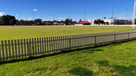 a smooth camera movement transitions from a white picket fence to a wide, empty sports ground under bright daylight, revealing clear skies and open space