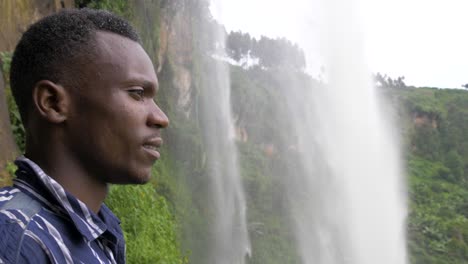 close up slow motion shot of a young african mans face as he looks out behind a tropical waterfall