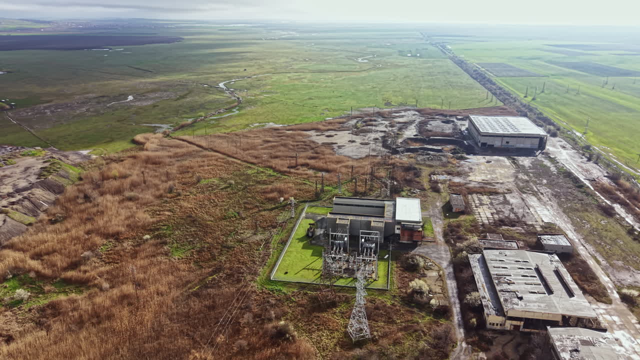 Old factory site with overgrown fields and power lines near a river in the countryside during daylight hours