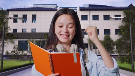 primer plano de una estudiante adolescente asiática con una mochila leyendo un libro y gritando una meta celebrando el éxito en el aprendizaje frente a un edificio escolar