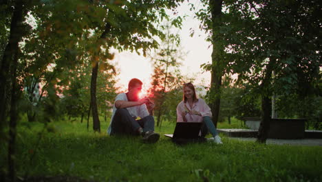 two people sitting outdoors with a laptop in front of them, toasting with bread slices in hands, enjoying nature, surrounded by trees with warm sunlight filtering through