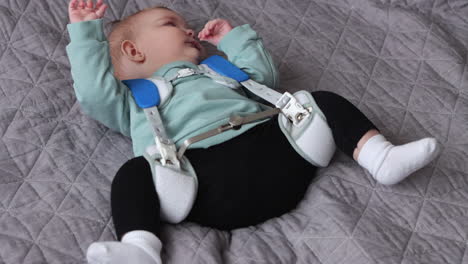 tired baby girl wearing pavlik harness to correct hip dysplasia lies on quilt