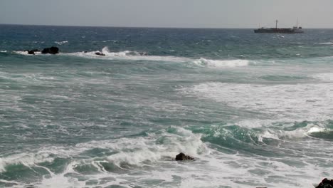 las olas se adentran en una costa rocosa con un carguero al fondo
