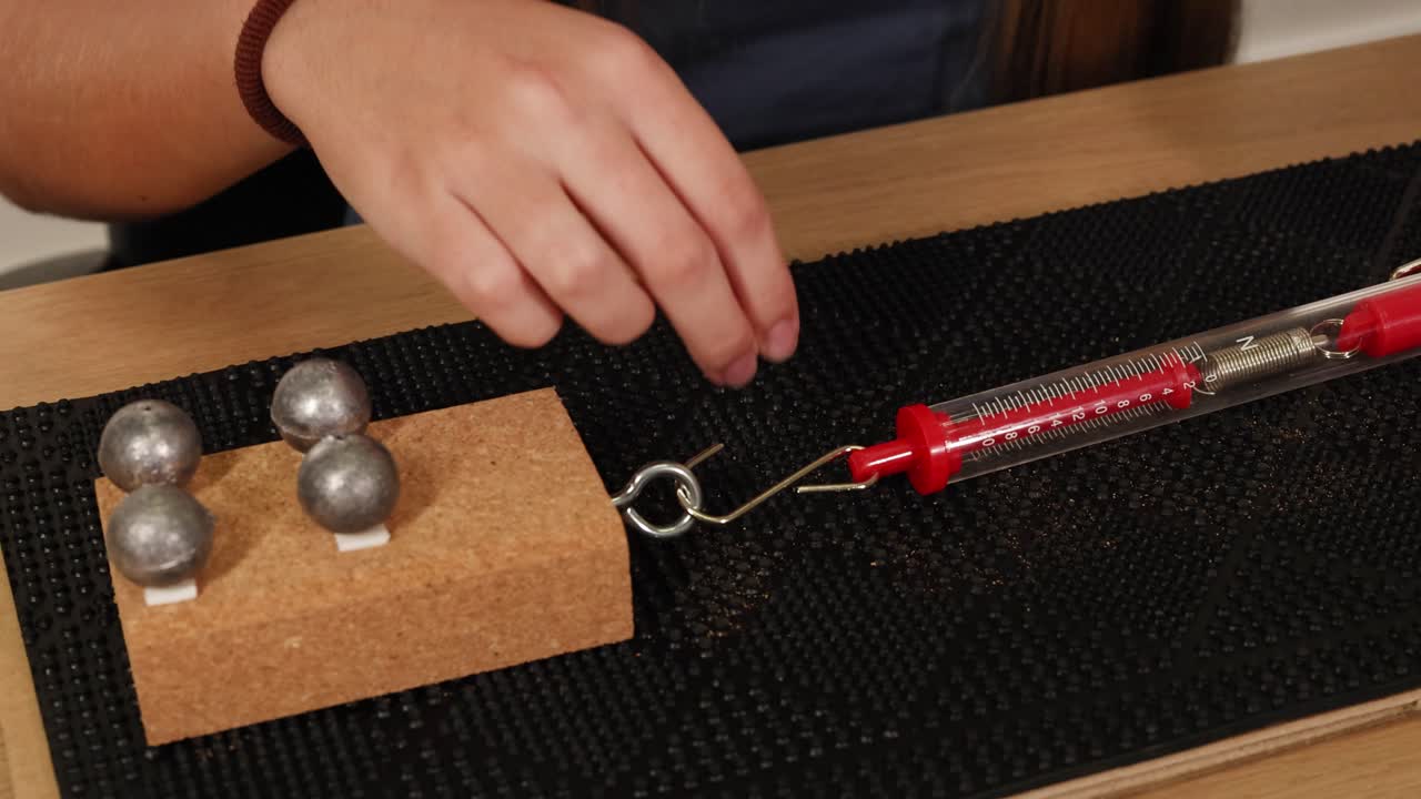 A hand uses a spring scale to measure the force needed to drag weights on a textured surface