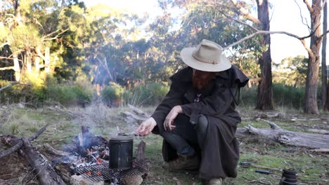 un bosquero en una chaqueta de cría de piel de aceite se sienta alrededor de un fuego con su billy hirviendo en el país alto