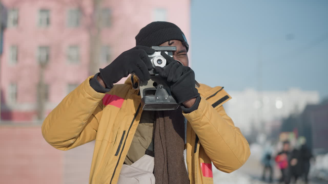 camera lifted up by lensman in yellow coat and gloves, one hand adjusts lens and snaps photo, reviews shot under clear blue sky on snowy city walkway with blurred buildings and passerby in background