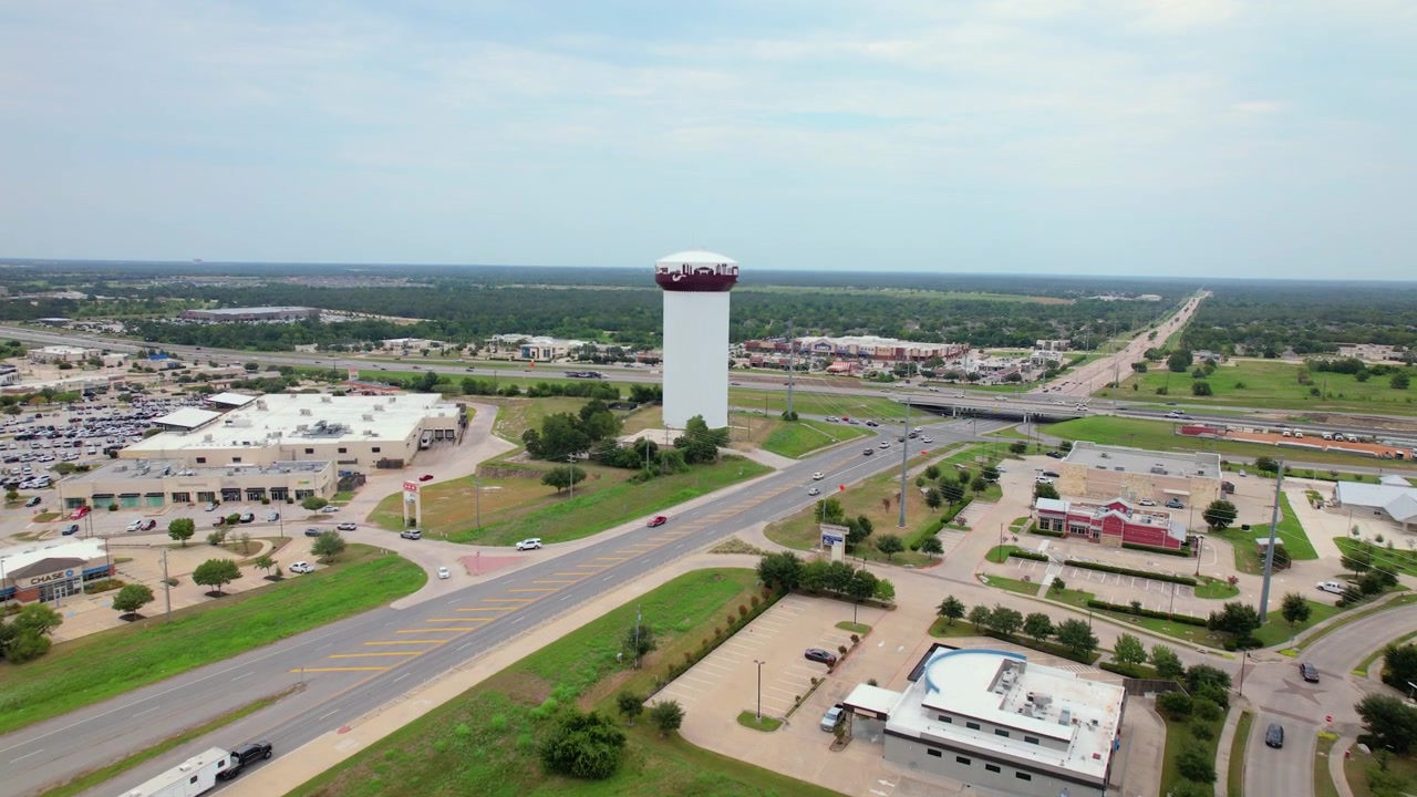 Riprese aeree con drone in volo in avanti da un'ampia inquadratura panoramica verso l'iconica torre idrica di College Station, con la scritta “Welcome to College Station”
