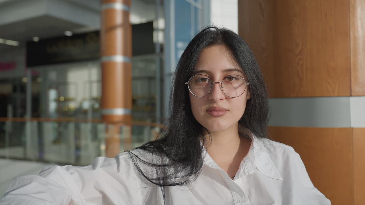 Confident young lady with long dark hair and eyeglasses rests against glass railing inside modern shopping mall, holding checkered coat and gazing peacefully with elegance and poise