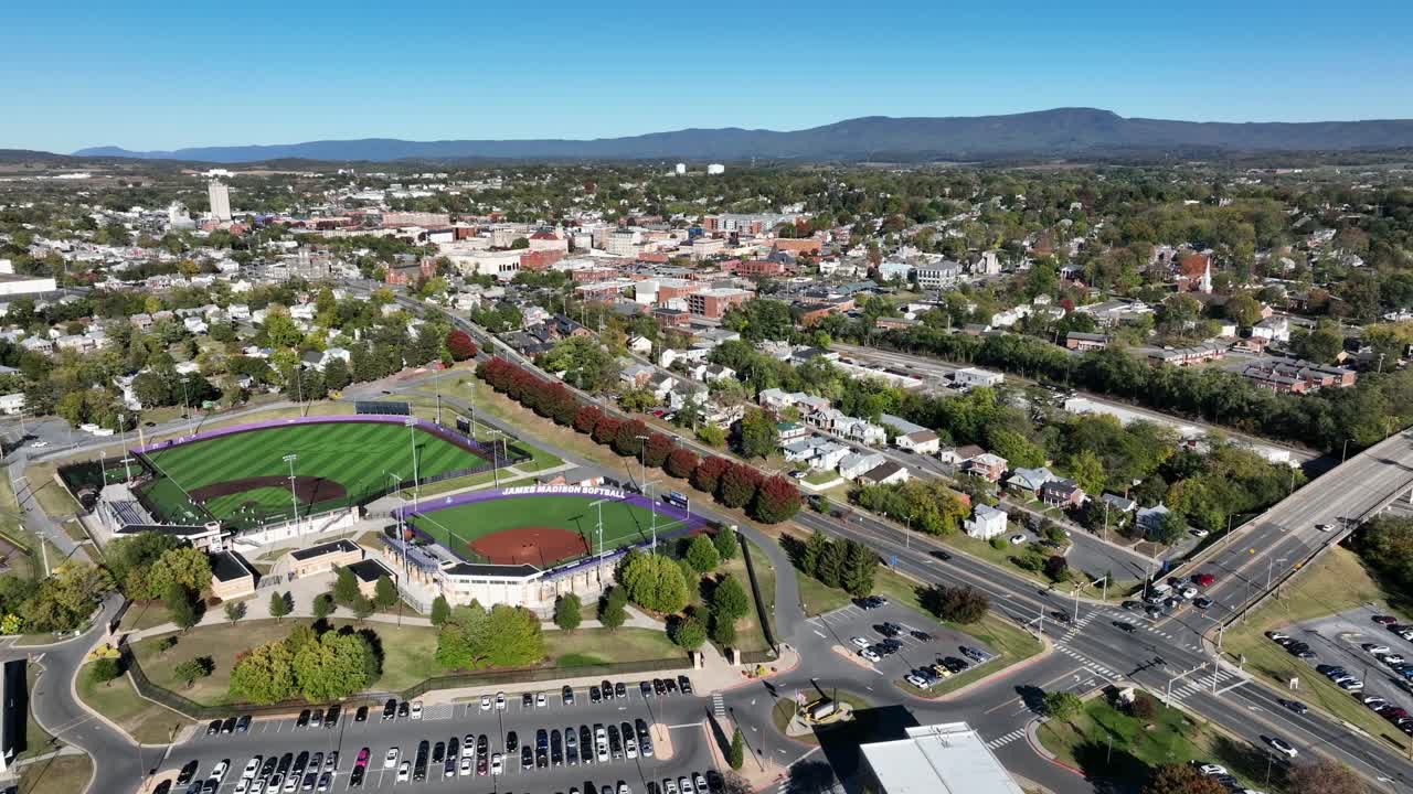 Drone flyover american baseball field stadium in Harrisonburg, Virginia. Colored trees and housing area with homes in suburbia of town. Downtown in background. Forward wide shot. Blue sky and sunlight