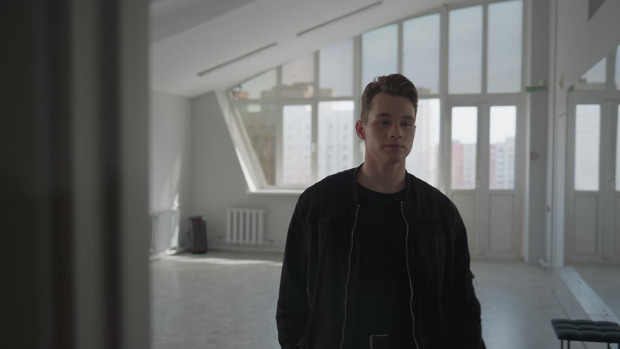 Fair skinned man in black jacket pulls wheeled travel bag into bright room with large windows overlooking high rise office building, concrete floor and radiator under soft natural light