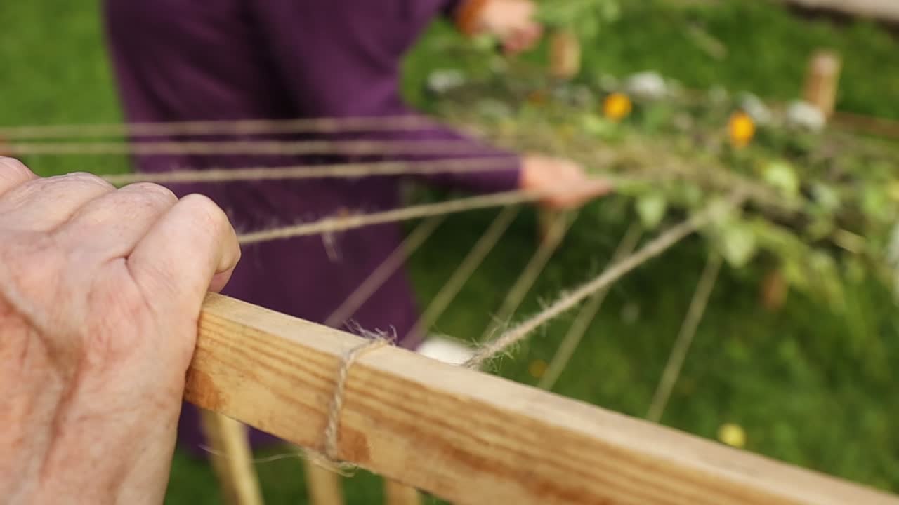women, one in Latvian folk costume, weave herbs, leaves, and flowers into an outdoor loom made of wooden stakes and string, beside a pond in a green garden