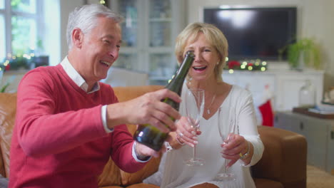 Amorosa-Pareja-De-Ancianos-Celebrando-Con-Champán-Alrededor-Del-árbol-De-Navidad-En-Casa