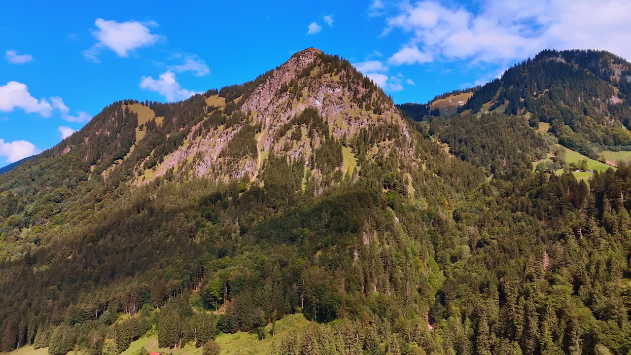 Approaching high rocks overgrown with pine tree woods. View of the Bavarian Alps at the backdrop of blue sky.