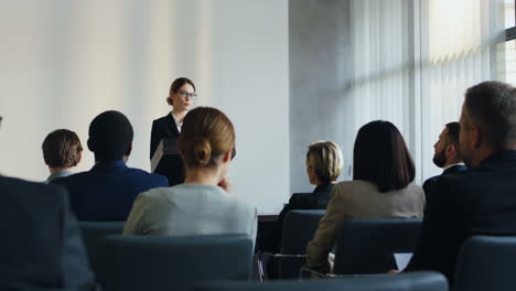 caucasian businesswoman speaker on a podium wearing formal clothes and talking in a conference room in front to many people