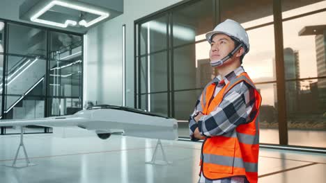 vista lateral de un ingeniero masculino asiático con casco de seguridad cruzando los brazos y mirando a su alrededor mientras está de pie con el proyecto de drones