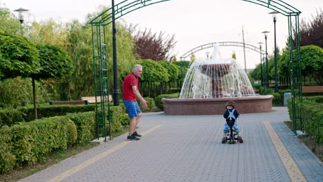 caucasian toddler riding his bicycle in the park. cheerful grandpa supporting a baby boy.