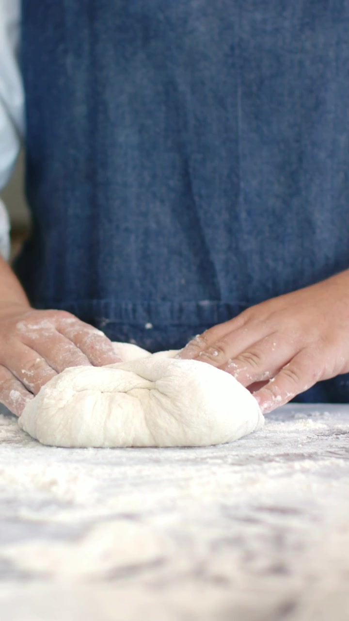 Vertical video: Floured hands shaping dough ball after resting at bench for proofing, denim apron