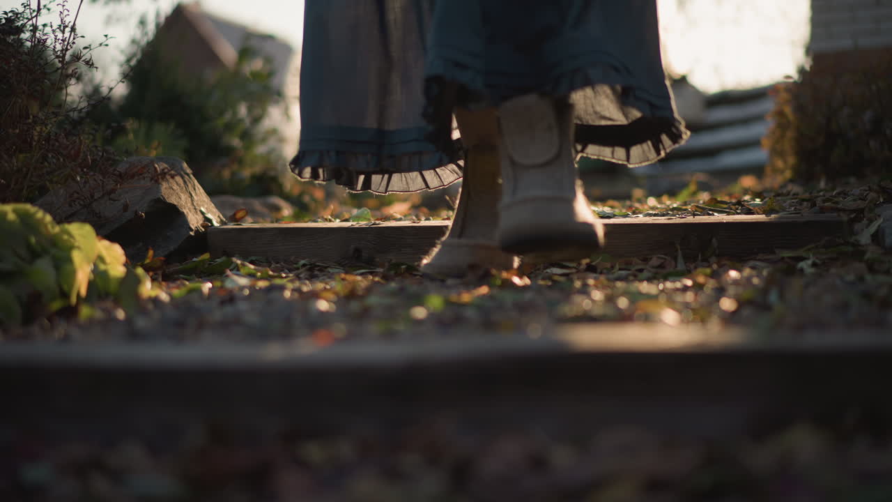 Low shot of boots stepping along garden path, long skirt and dried bouquet visible in warm late light, gravel and fallen leaves crunch underfoot, intimate detail of harvest return toward home kitchen