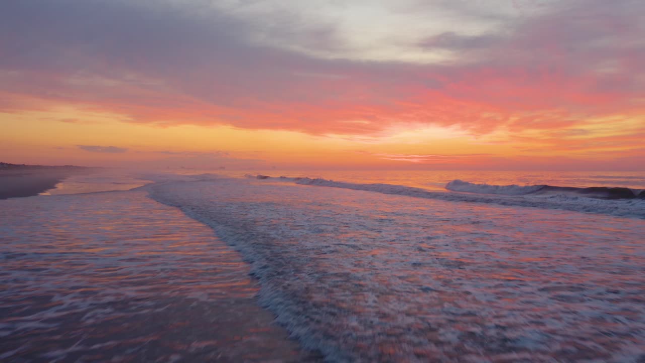 Low to high aerial over small breaking waves, purple and orange sunset in background